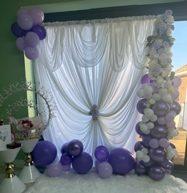 A decorative setting with white drapes, purple balloons, and floral accents.